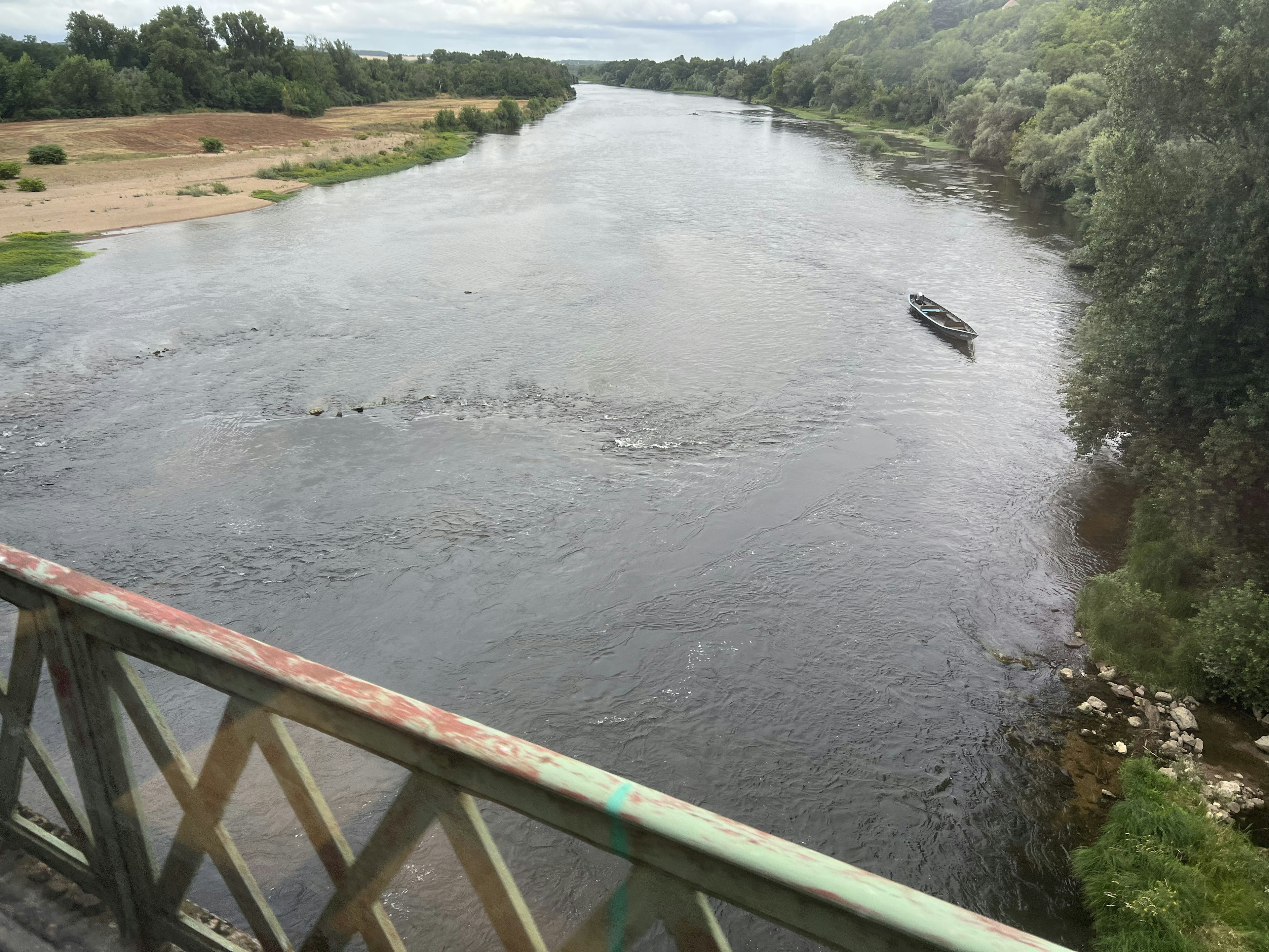 Picture of a river surrounded with trees and sand, taken from a bridge. A small boat is floating on it.